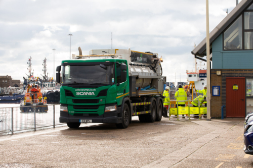 A green Willow Pumps tanker parked in front of a dock with Willow Pumps workers behind a barrier at the back of the tanker.