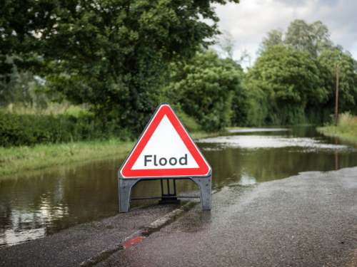 A red and white "Flood" sign in front of a large puddle across a country road.